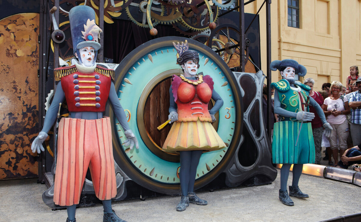Drei kostümierte Pantomimenspieler auf dem Gauklerfest stehen vor einer großen Uhr und schauen ins Publikum. ©© GDKE, Ulrich Pfeuffer