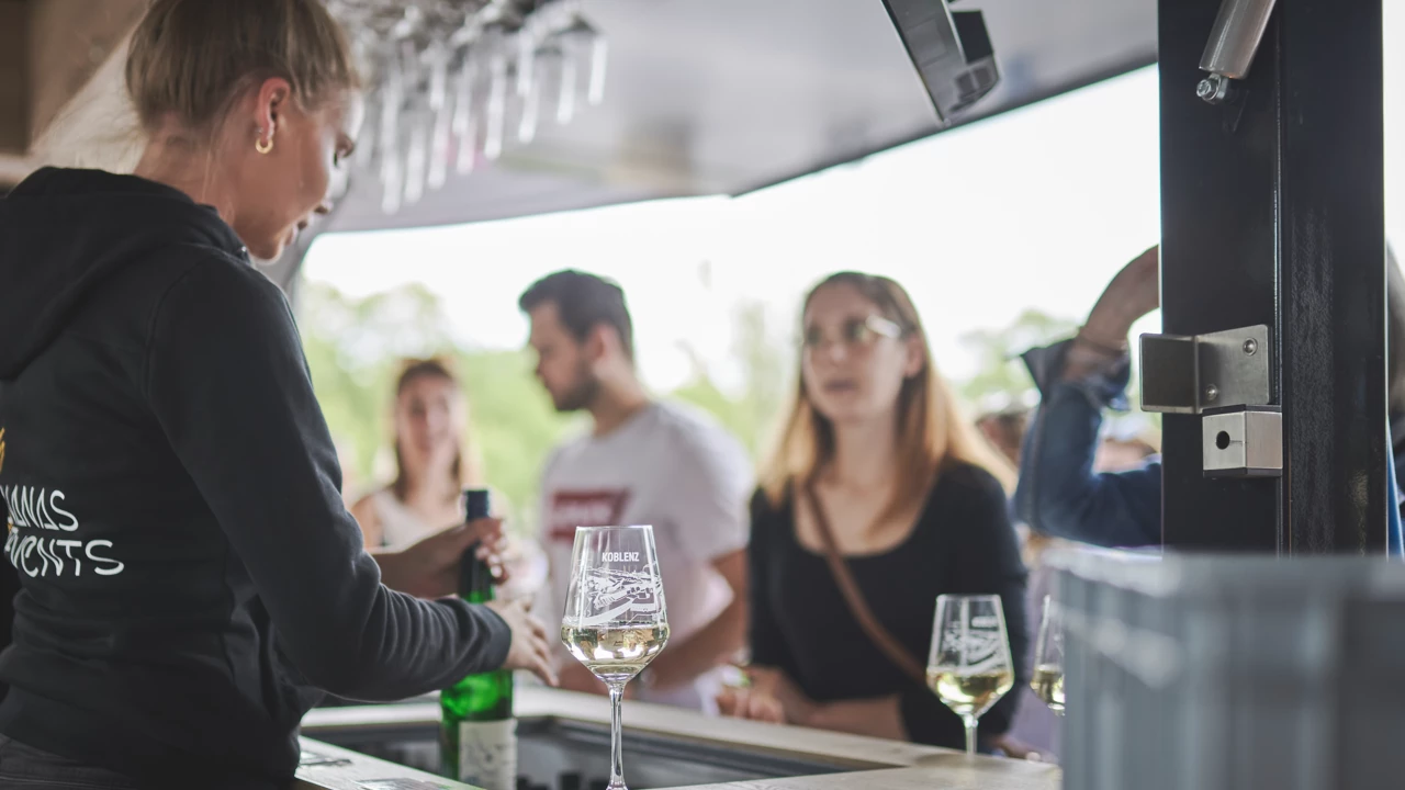 Ein Glas Wein steht auf der Weintheke und Kunden bestellen