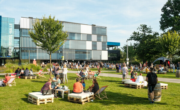 Menschengruppen sitzen verteilt bei der Blaue Stunde vor der Rhein-Mosel-Halle ©Camaze Menschengruppen sitzen verteilt bei der Blaue Stunde vor der Rhein-Mosel-Halle ©Camaze