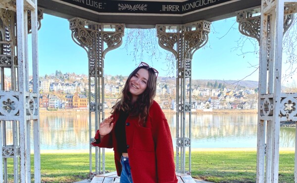 Frau mit rotem Mantel und Sonnenbrille steht im Augusta-Pavillon in den Rheinanlagen, im Hintergrund der Rhein und der Stadtteil Pfaffendorf. ©Kaya Reichert Frau mit rotem Mantel und Sonnenbrille steht im Augusta-Pavillon in den Rheinanlagen, im Hintergrund der Rhein und der Stadtteil Pfaffendorf. ©Kaya Reichert